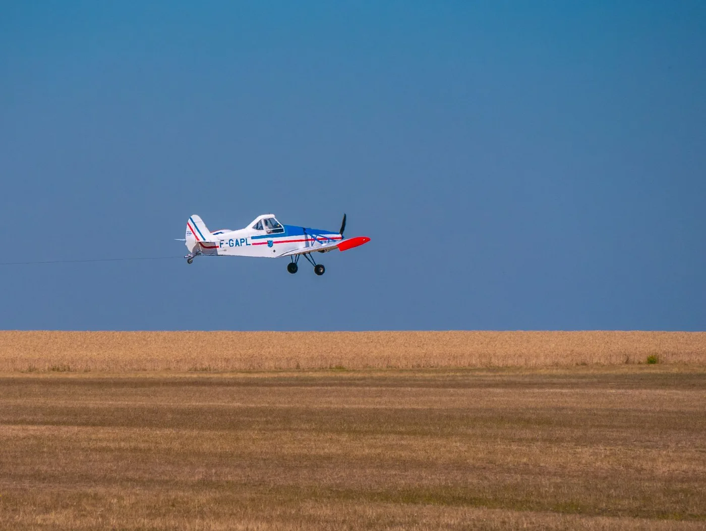 Small agricultural airplane flying low over a golden wheat field under a clear blue sky