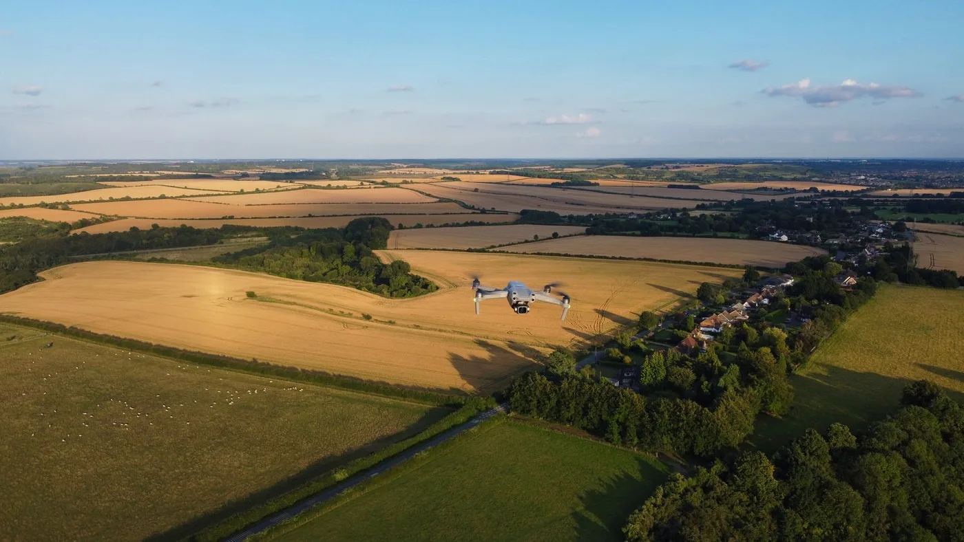 Small rotary-wing mapping drone hovering low over golden wheat fields at sunset with a village in the distance